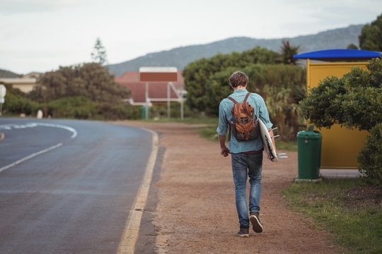 Man with backpack carrying a surfboard walking along road