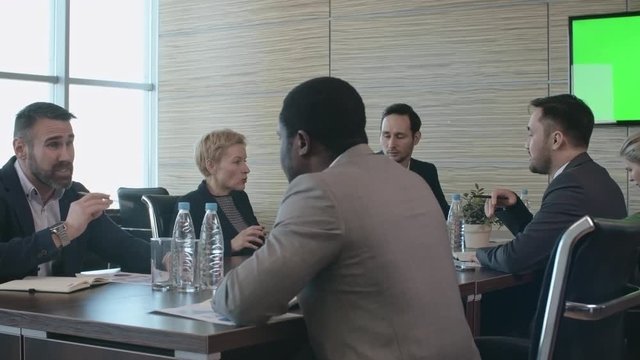 Pan of bearded man sitting behind table in conference room and discussing finances with African businessman
