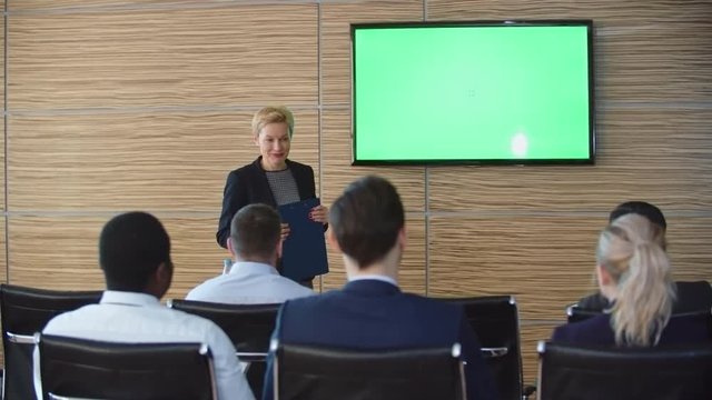 Crane shot with tilt up of blond businesswoman giving presentation before audience in conference room, LED TV with green chroma key in background