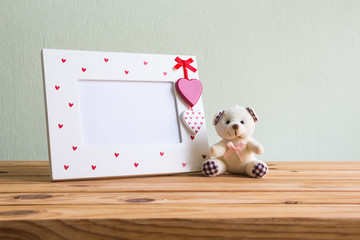 White vintage photo frame with red heart and teddy bear on wooden table over wall grunge background, Valentine day concept.