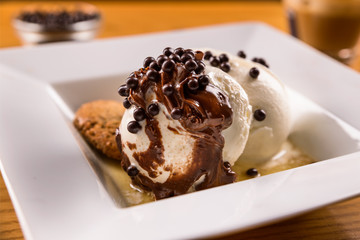 Vanilla ice cream balls in a white plate with cookies and sweet sauce covered with chocolate and chocolate balls on a wooden plate.