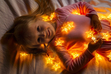 little girl lying on the bed in the bedroom and wrapped in Christmas lights in the shape of stars warm yellow