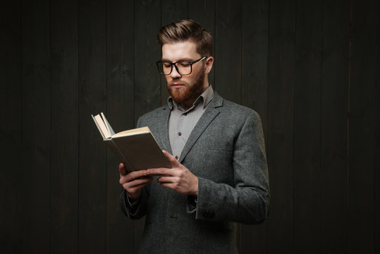 Thoughtful young man in eyeglasses standing and reading book