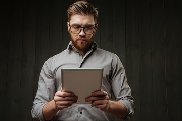 Concetrated bearded man in eyeglasses and shirt using tablet computer