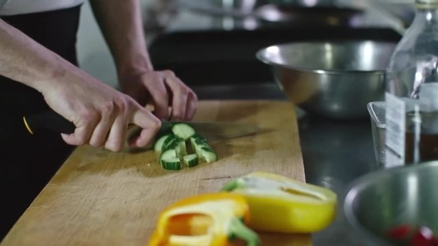 Closeup of hands of man dicing cucumber on wooden board and then putting pieces into metal bowl