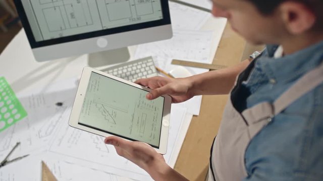 Handheld high angle shot of male artisan comparing his blueprints in workshop