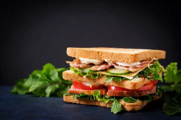 Fotobehang Grote Clubsandwich met ham, bacon, tomaat, komkommer, kaas, eieren en kruiden op donkere achtergrond © timolina