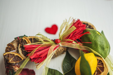 Плетеный венок в виде сердца, украшенный листьями, лимонами, сушеным лимоном, бантами, божьей коровкой, лепестками роз на белом деревянном столе