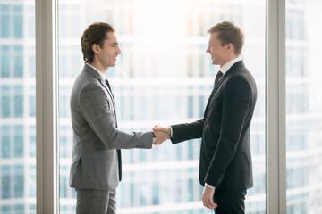Fototapeta premium Side view portrait of two smiling business partners making agreement, taking a big deal and shaking hands in meeting room at office. Urban style glass office building in big city on the background