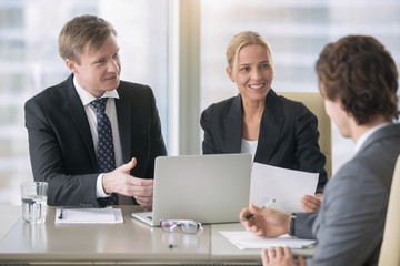 Group of three business partners discussing new project at meeting in office room, using laptop. Businesspeople in formal wear suits interacting, brainstorming. City office building on the background