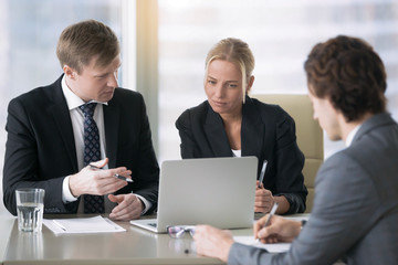 Fototapeta premium Group of three business partners discussing new project at meeting in office room, using laptop. Businesspeople in formal wear suits interacting, brainstorming, looking presentation on laptop screen