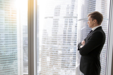 Fototapeta premium Successful businessman in classical formal suit standing in office with hands crossed on chest, looking through window at big city skyscrapers, thinking of new deals, dreaming. Copy space for text