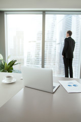 Company president in formal suit standing, looking through window at cityscape, dreaming, planning new projects, resting after intensive work. Focus on laptop on office table. Business success concept