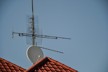 TV antennas and satellite dish for television mounted on the tiled roof of house isolated on blue sky background in countryside. Telecommunications aerials and receiver devices on rooftop of building © lukasbeno
