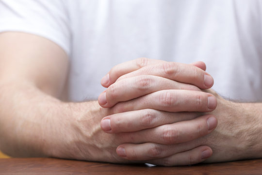 Man is sitting and holding his hands together on a wooden table. Like praying or in a meeting listening.