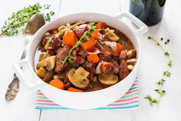 Beef Bourguignon in a casserole on white table. Stewed with bacon, garlic, carrots, onions, mushrooms,  and spices. Bunch of fresh thyme, vintage spoon, red wine.