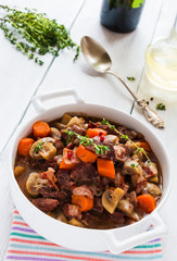 Beef Bourguignon in a casserole on white table. Stewed with bacon, garlic, carrots, onions, mushrooms,  and spices. Bunch of fresh thyme, vintage spoon, red wine.