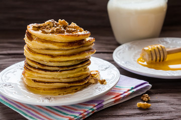 Homemade pancakes with honey and walnuts, vintage white plate, dipper, milk in glass jar, dark wooden table.
