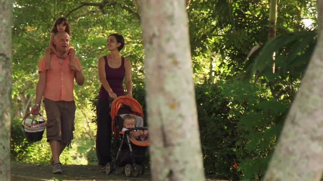 Happy couple with son and daughter walking in park. Family with man, woman and children doing picnic, having fun outdoor. Joy for husband, wife, newborn child in stroller and little girl. Slow motion