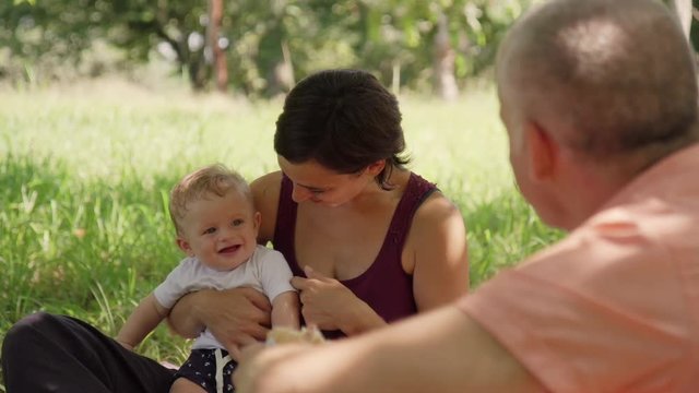 Happy white people with infant doing picnic in city park. Caucasian family with man, woman and child eating, having fun outdoor. Leisure for dad, mom, baby boy. Slow motion