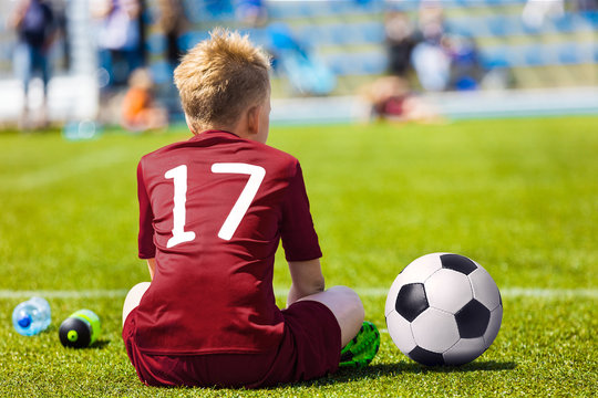Young Soccer Football Player. Little Boy Sitting on Soccer Pitch. Youth Football Player in Red Soccer Jersey
