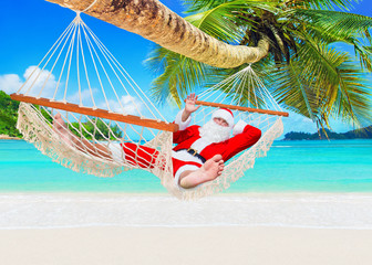 Positive smiling Santa Claus relax in white cozy mesh hammock under coconut palm tree at exotic island sandy ocean beach - Happy New Year and Merry Christmas travel destinations, welcome hand gesture