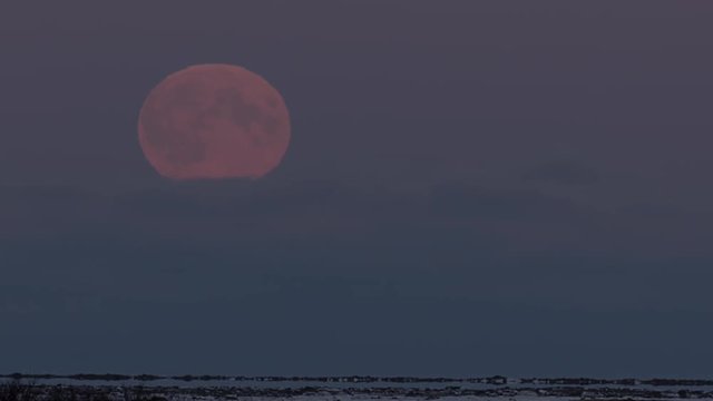 Full moon rises behind clouds over frozen tundra at dusk