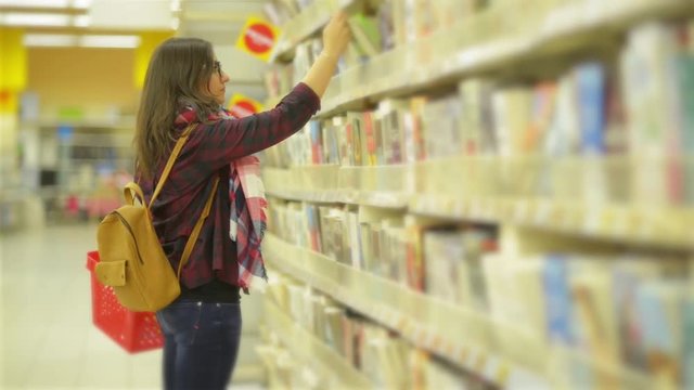 Young Beautiful Girl Wearing Scarf and Glasses Choosing a Book in the Supermarket. Woman with Long Dark Hair and Yellow Backpack Takes a Book and Browses a Pages