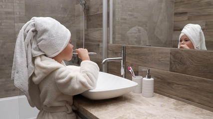 Cute little girl in a bath towel on the head and a bathrobe cleaning teeth in the bathroom against...