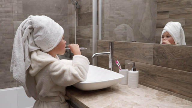 Cute little girl in a bath towel on the head and a bathrobe cleaning teeth in the bathroom against the mirror
