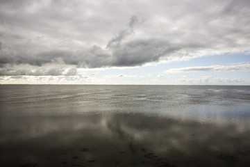 shoreline of Ameland Island, with view over the wadden sea, with clouds and tourmented sky reflecting in water at dawn