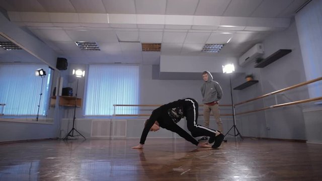 Two young hip-hop dancers participating in dance battle. Young man in grey pullover and trousers watching his friend performing urban dance in the classroom.