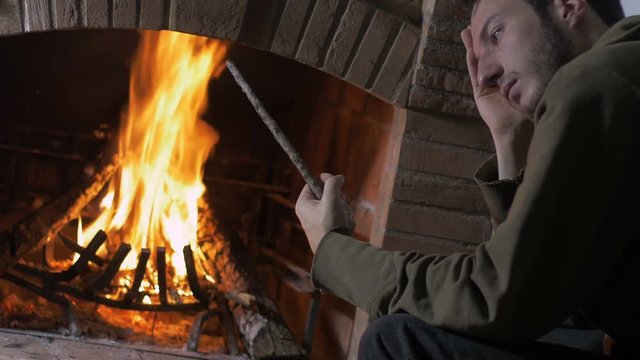 young man sat at home near a fire place, plays pensively with a stick