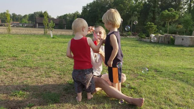 A mom blow bubbles, while her two sons stand nearby in fascination.  They pop some bubbles with their hands. 4k. Slow motion.