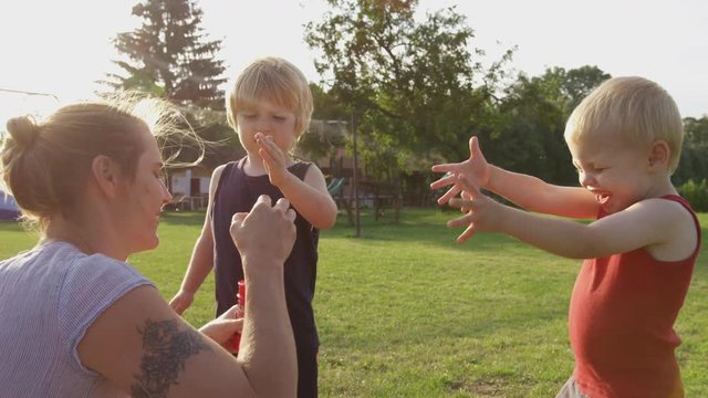 A mom blows bubbles into the breeze with her two sons standing nearby.  Some bubbles pop on the face of one son, who reacts with surprise. 4k. Slow motion.