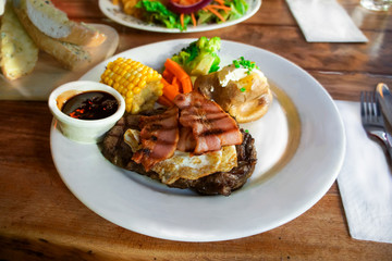 Plate of delicious piece of well done stake with fried egg and bacon on top. Serve with BBQ sauce, corn, green vegetable and bake potato. Garlic cheese bread on the side