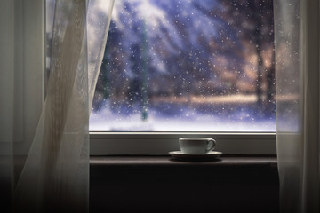 Cup on the windowsill. Outside, winter fairy tale - the evening, a beautiful snow falls. The concept of comfort in the home, loneliness, winter evening