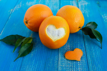 A few ripe oranges with leaves on a blue wooden background. One cut orange peel in the shape of a heart.