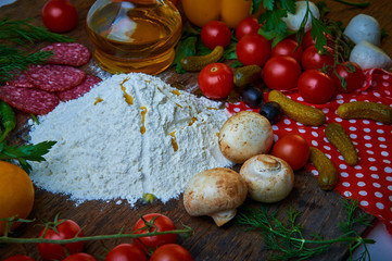 Various ingredients for pizza. Flour, butter, mushrooms, cucumbers, cherry tomatoes, olives,Mozzarella  , sausage, cheese. Dark background.