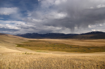 Fototapeta na wymiar A highland swamp river valley with yellow grass on a background of snow covered high mountains and glaciers under clouds and blue sky, Plateau Ukok, Altai Siberia, Russia