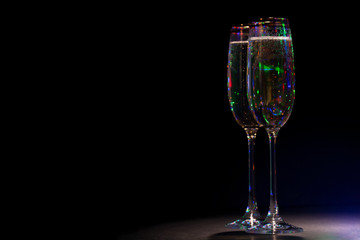 Two glasses with champagne on a dark background. The glass reflects colored lights.