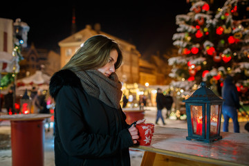 Long hair girl on European Christmas Market. Young woman Enjoying Winter Holiday Season. Blurred  Lights background, dusk. Cups with drink in hand. Selective focus