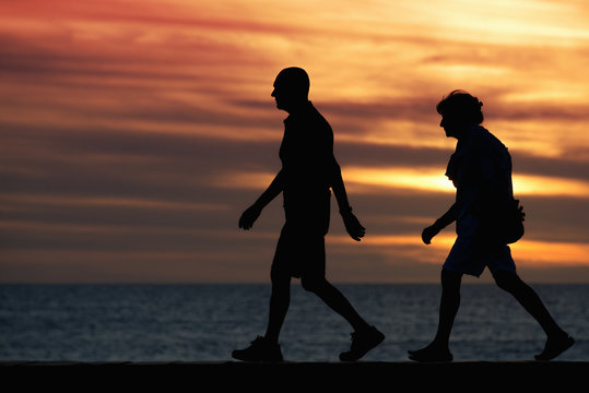 Seniors Walking Brisk Walk Along The Shore At Sunset