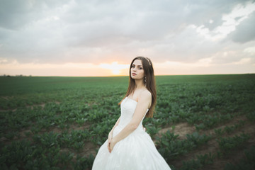 Fototapeta na wymiar Beautiful brunette bride in elegant white dress holding bouquet posing neat trees