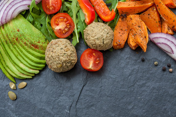 Fototapeta na wymiar Vegan food frame: avocado, sweet potato, lentil cutlets, tomatoes, arugula, onions on black stone background with a copy space in the center