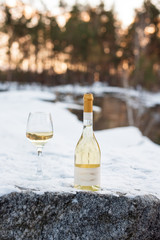 Love, romance, holiday, New Year celebration concept. Bottle and glass of white wine chilled by snow in winter forest on sunset.