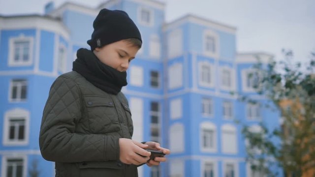 In the courtyard stands a boy on footpath. He wore a jacket, a black hat and a scarf around his neck. He played on your phone. Against the backdrop of a blue house