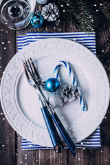 Christmas table setting. White plate, knife and fork, napkin. Silver and Blue Christmas Decorations, fir branch, candy cane on wooden background table. Top view.