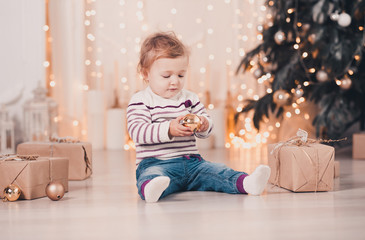 Cute baby girl under 1 year old holding Christmas ball sitting on floor over Christmas tree with presents in room. Childhood. Celebration.