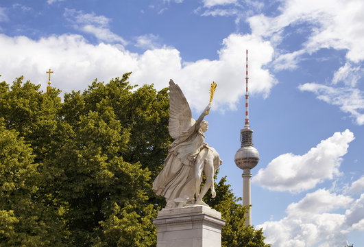Winged woman statue on Schlossbruecke bridge with trees, cloudy sky and TV tower in the background. Designed by K. F. Schinkel in 1821 with sculptures of gods.